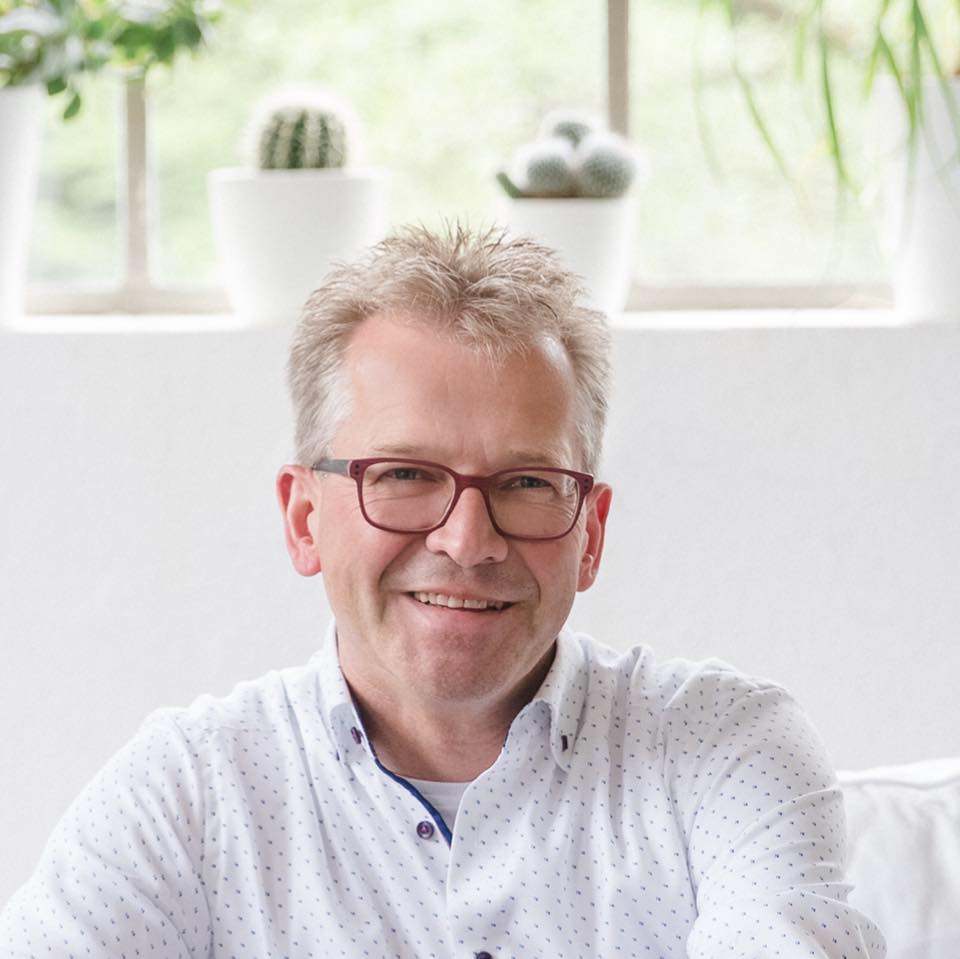 Porträt eines lächelnden Mannes mit Brille und hellem Hemd in einem hellen Innenraum.