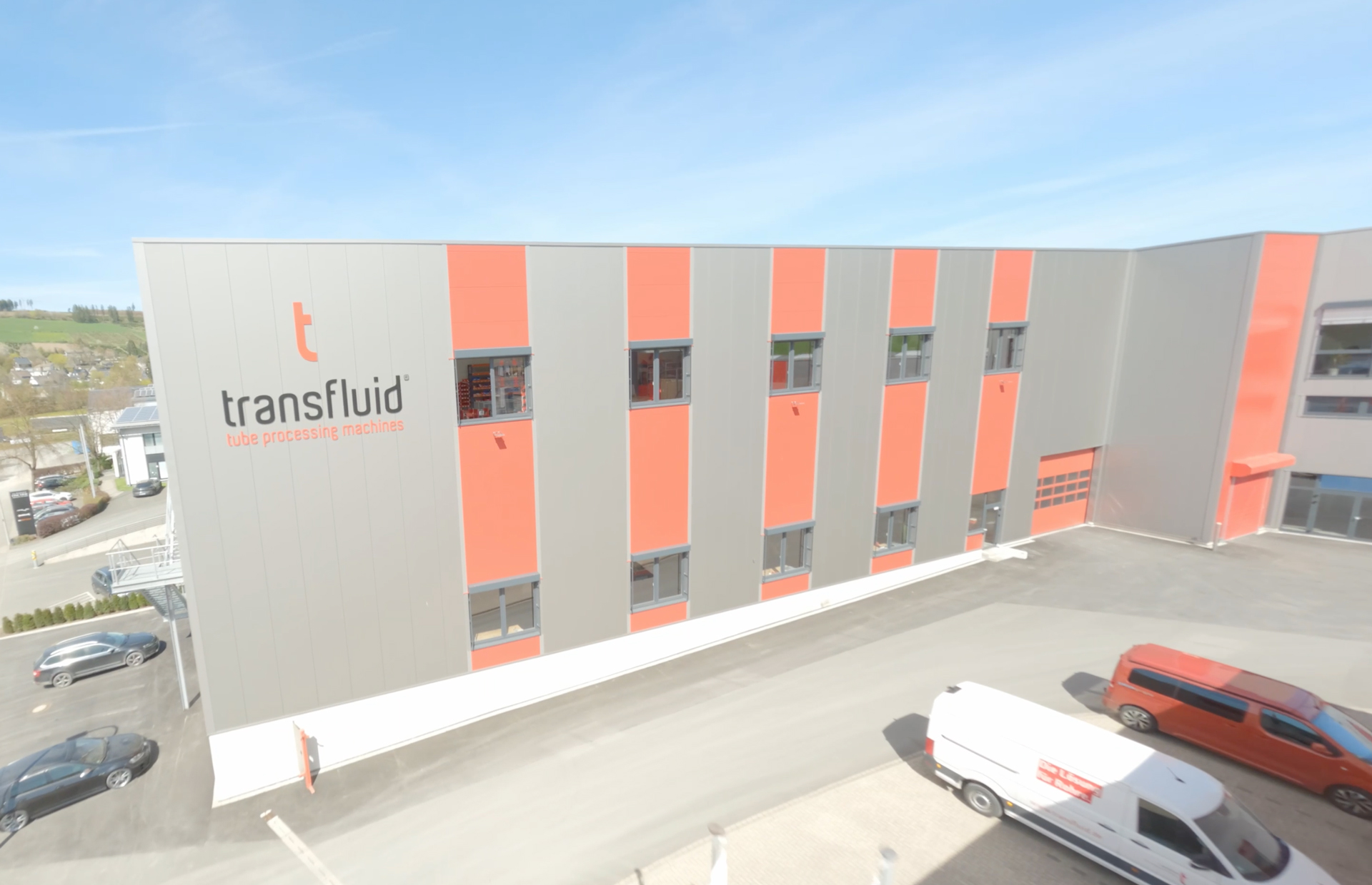 Moderne Firmenhalle von transfluid mit grau-roter Fassade und Firmenlogo „transfluid – tube processing machines“ bei sonnigem Wetter in Schmallenberg.