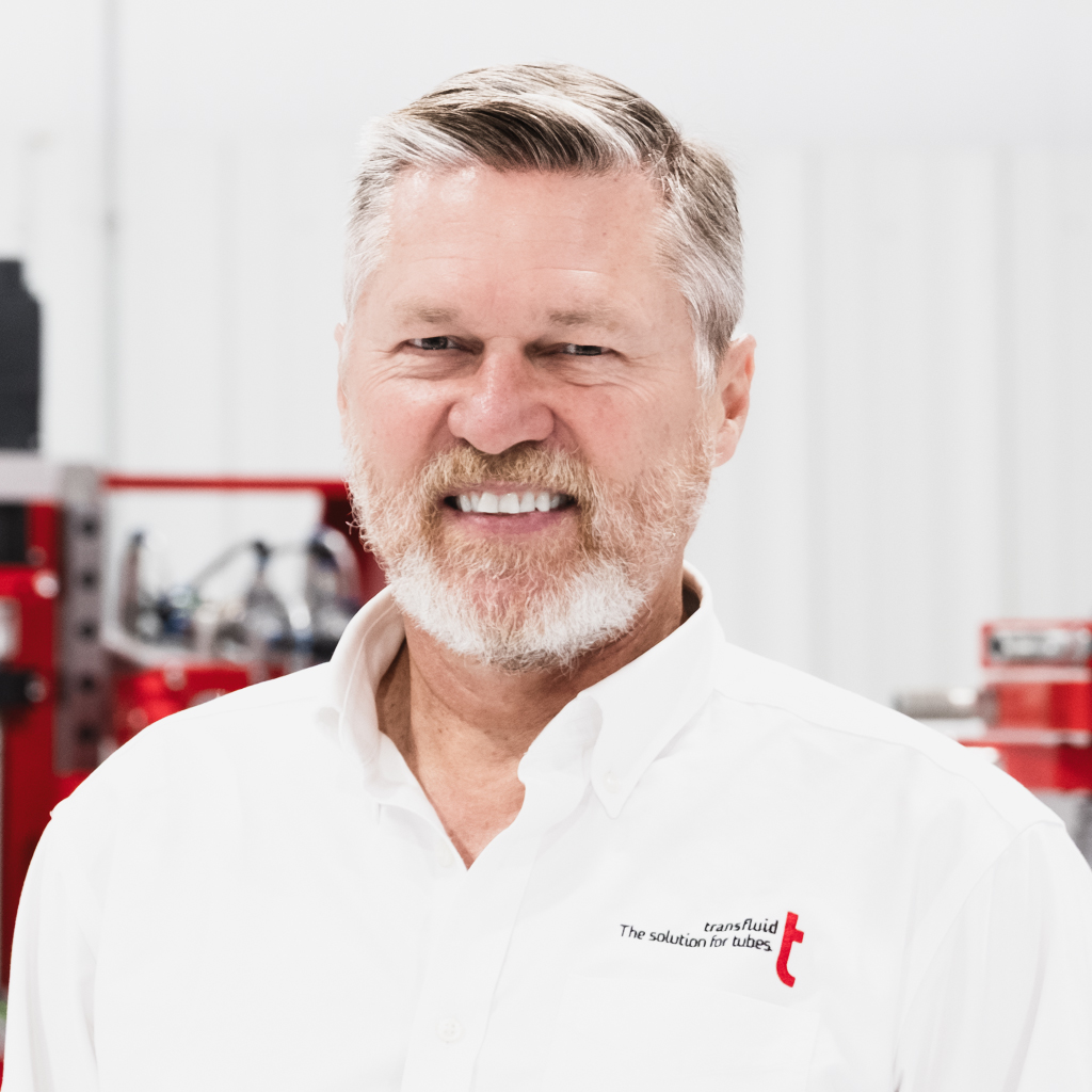 Mitarbeiter mit grauem Haar und Bart im weißen Firmenhemd vor Industrieanlage.