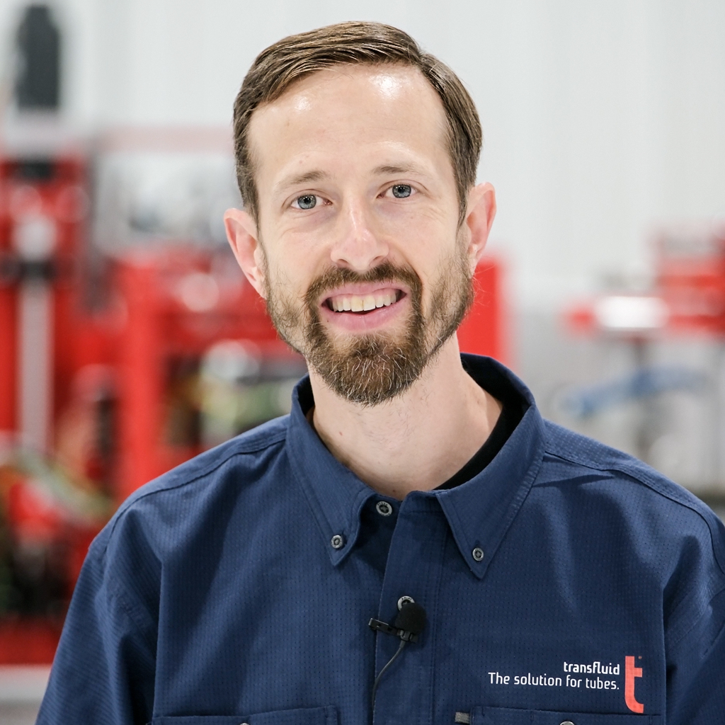 Mitarbeiter mit Bart im blauen Firmenhemd mit Mikrofon vor Industrieanlage.