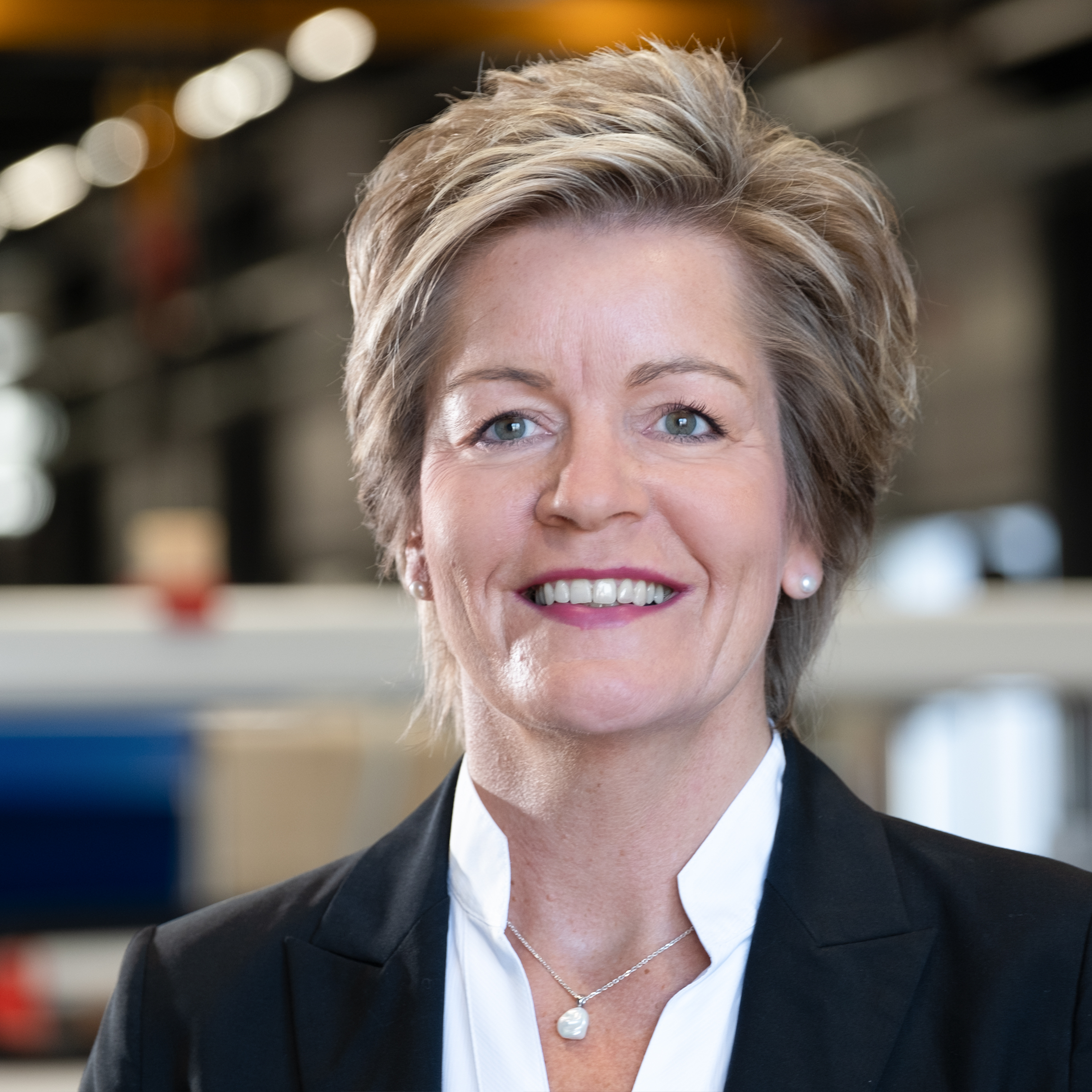 Mitarbeiterin im Blazer in moderner Industriehalle mit unscharfem Maschinenhintergrund.